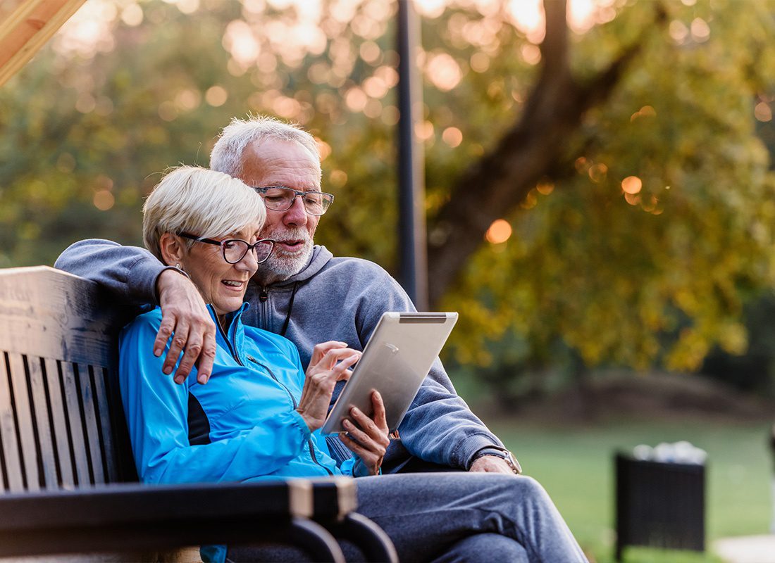 Medicare - Senior Couple Smiling Together While Looking at a Tablet on a Park Bench