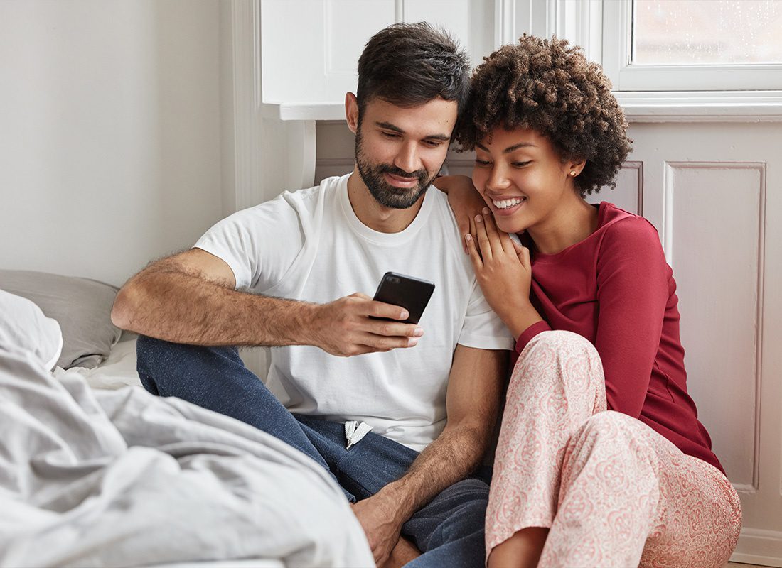 Read Our Reviews - A Couple Smiling While Looking at A Smartphone in Their Pajamas